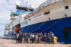 Twenty-one members of an expedition team pose for a photo in front of a research ship. The research ship has a blue-and-white hull with the name Falkor (too) printed on it. In the background is cloudy blue sky.