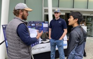 Two engineers talk to an aquarium visitor in front of a table outdoors in front of a building entrance. The engineer on the right is wearing a gray cap, a gray vest, and a blue sweatshirt. The engineer on the right is wearing a blue cap, navy blue polo shirt, and blue jeans. The visitor is wearing a black-and-blue jacket. The table has a banner that reads FathomNet with several scientific posters. In the background is the entrance to a public aquarium with a sliding glass door and frosted white windows.