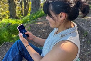 A person sitting in a park plays the FathomVerse on their mobile phone. The person has shoulder-length black hair in a ponytail and is wearing a sleeveless sweater with beige and white stripes and blue jeans. They are holding a smartphone in a pink case. The person is outdoors and in the background on the right are terraces of brown dirt, and on the left are green grass and brown tree trunks.