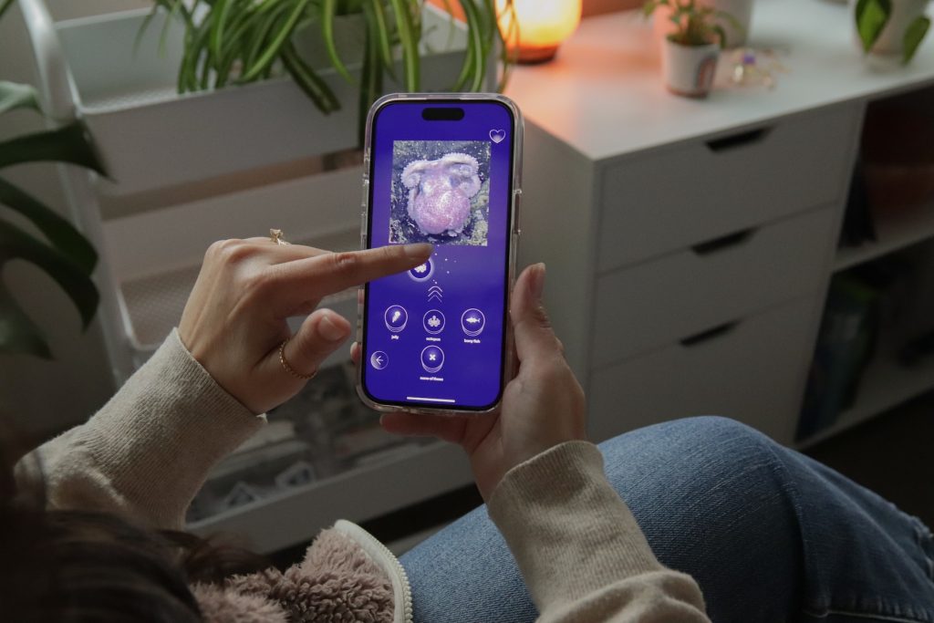 A person wearing a tan sweater and blue jeans is seated on a couch holding a smartphone while playing the FathomVerse game. The gamer is dragging a digital label across the phone screen to tag a photo of a purple octopus. In the background is a home office with white cabinets and green plants.