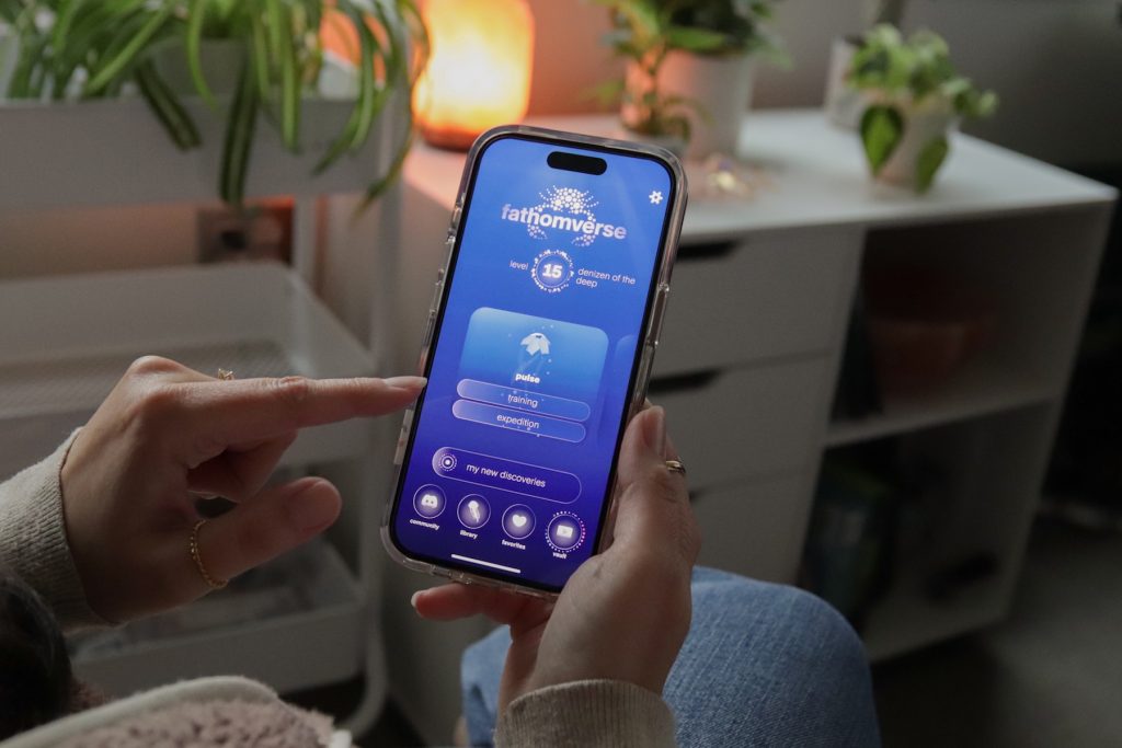 A person wearing a tan sweater and blue jeans sits on a couch holding a smartphone. She is tapping on the home screen of the FathomVerse mobile game. In the background is a home office with white cabinets and green plants.