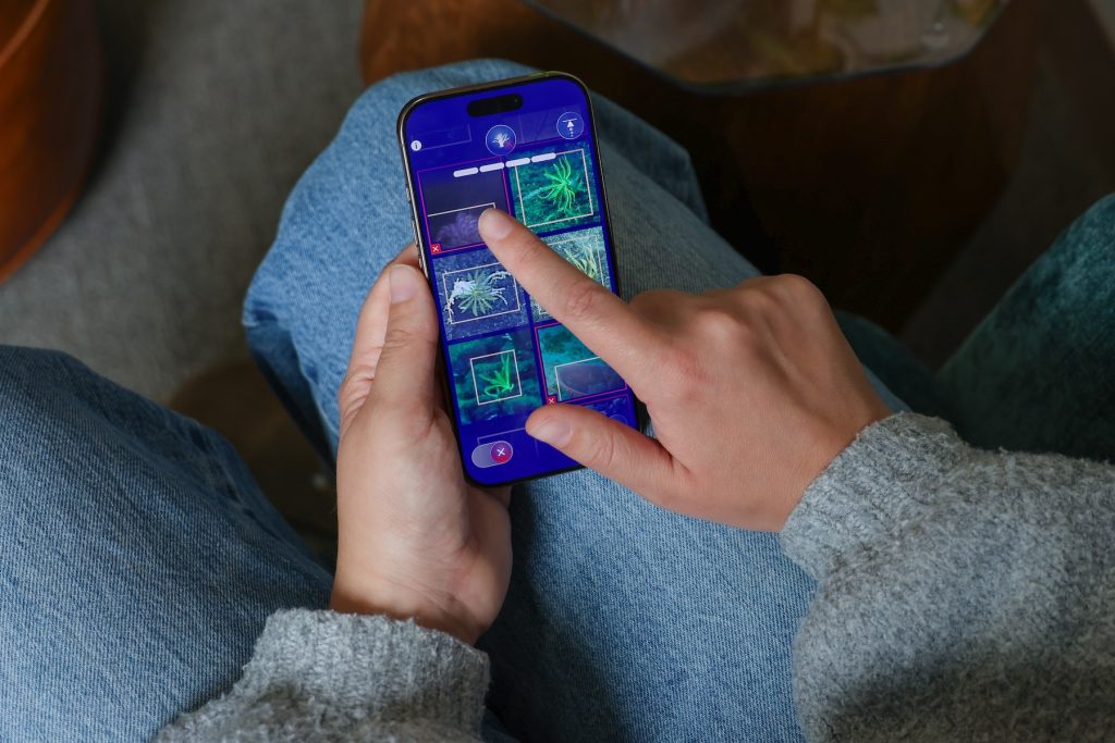A person seated in a blue armchair plays a mini-game in the FathomVerse app on her iPhone. She is wearing a gray sweater and blue jeans. On her phone screen is a grid of six photos on a blue background with app icons. She is tapping one of the photos to add a red box. In the background is gray carpet.