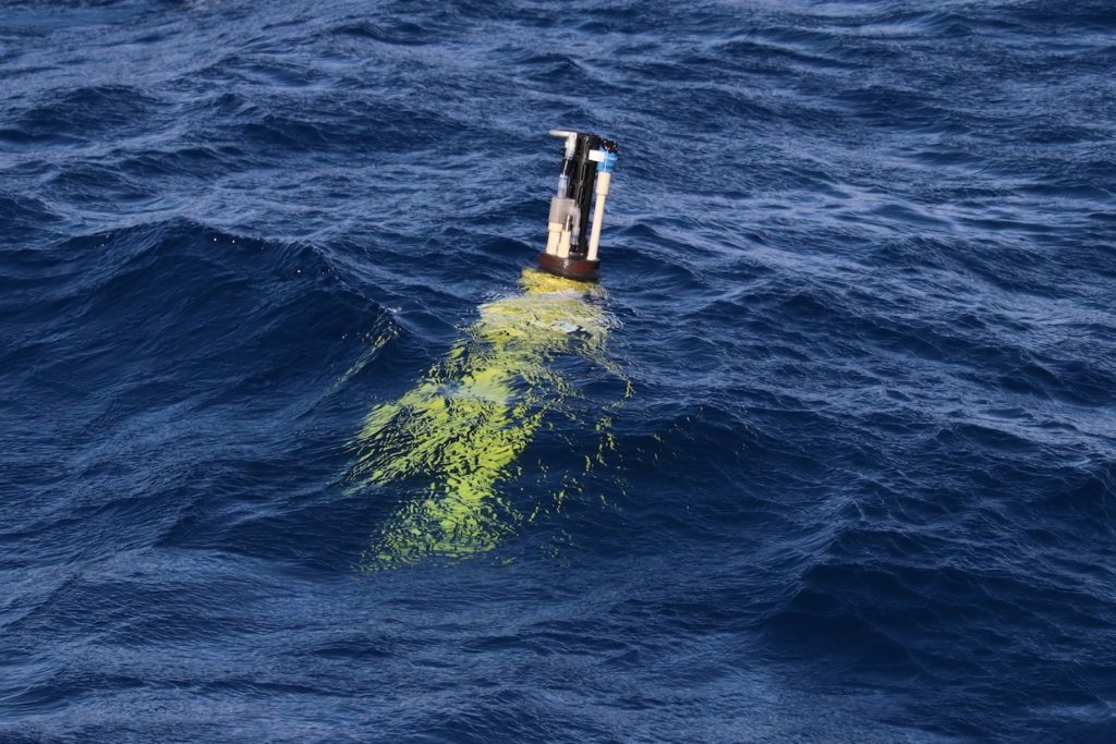 A robotic float drifts at the ocean surface. The float has a yellow plastic housing. At the top are white and black sensors protruding out of the water. The background is blue ocean.