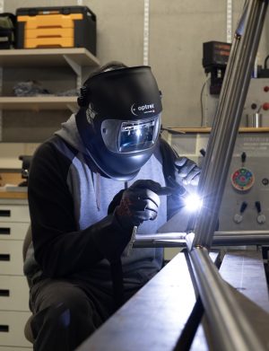 An MBARI mechanical technician welds the metal frame of a scientific instrument. The technician is wearing a black welding helmet, gray sweatshirt, and black pants. In the background is a machine shop workbench.