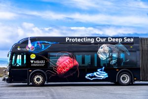 The front of a large black bus parked along the Santa Cruz coastline. The bus features large images of animals, including a transparent squid, a red jelly, a white rope-like siphonophore, and a brown fish. The bus also has large white text reading “Protecting Our Deep Sea.” This photo shows the bus parked on dry gray asphalt with blue sky and wispy white clouds in the background.