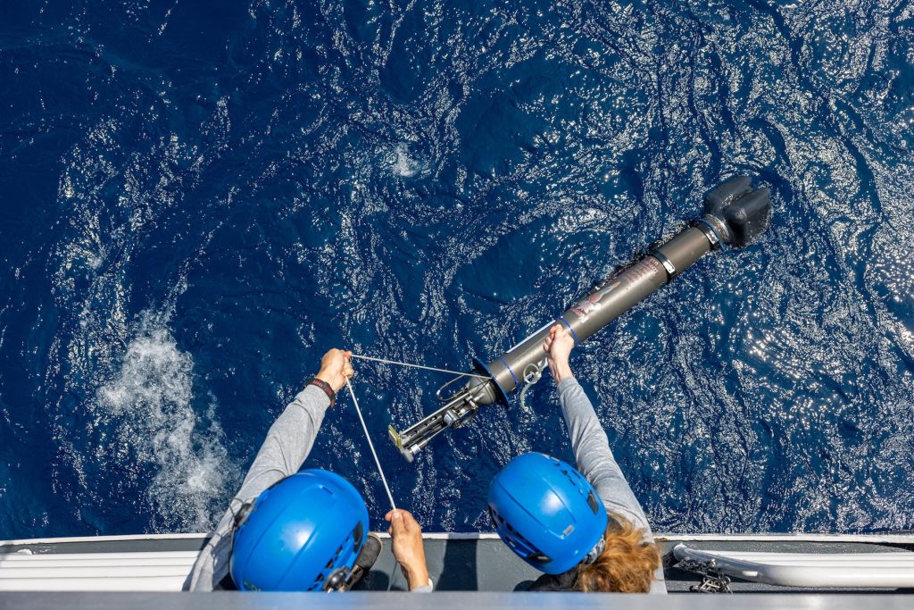 An overhead photo shows two researchers lowering a robotic float into the ocean. The researchers are wearing blue hard hats and gray jackets and leaning over the deck of a research ship. They are holding rope lines connected to a cylindrical float with a gray plastic housing. Below is blue ocean.