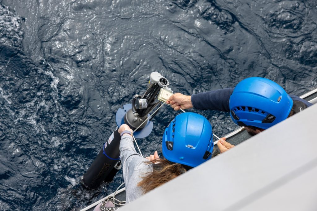 An overhead photo shows two researchers lowering a robotic float into the ocean. The researchers are wearing blue hard hats and leaning over the deck of a research ship. They are holding rope lines connected to a cylindrical float with a gray plastic housing. Below is blue ocean.