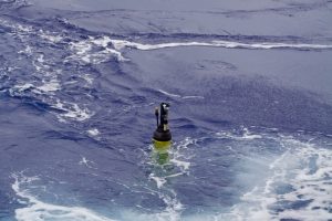 A robotic float drifts at the ocean’s surface. The float has a cylindrical yellow plastic housing underwater with black instruments at the top extending above the water’s surface. The background is blue ocean, with choppy white water in the foreground.