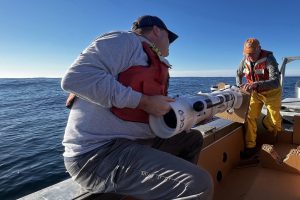Two researchers prepare to deploy a robotic float over the side of a small boat. The researcher in the foreground is wearing a blue cap, gray sweatshirt, orange life vest, and gray pants, and is sitting on the railing of the boat. The researcher in the background is wearing an orange cap, a gray shirt, an orange life vest, yellow waders, and brown boots, and is standing near the stern of the boat. They are each holding one end of a cylindrical robotic float with a white plastic casing. The foreground is brown cardboard laid out on the deck of a boat. In the background are blue ocean, blue sky, and gray fog on the horizon.