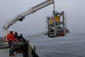 Two members of MBARI’s marine operations crew deploy a scientific instrument over the side of a research ship. The crew are wearing white hard hats and orange life vests. The instrument has a silver metal frame with yellow floats at the top and two silver metal spheres on a green fiberglass grate at the base. The instrument is suspended from a white metal crane at the top of the frame, and the crew are guiding the instrument into the water with ropes. On the left side of the frame is the silver metal deck of a research ship. On the right side of the frame is gray ocean, with overcast gray sky on the horizon.
