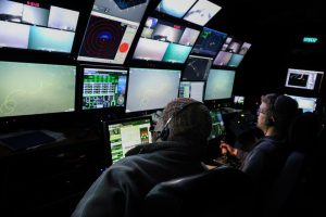 Two submersible pilots wearing black microphone headsets operate a deep-sea submersible from a dark control room aboard a research ship. The pilots are seated in large chairs with their backs to the camera. They are watching a wall of monitors displaying video and data feeds from the submersible. The pilot on the right has his right hand on a joystick control.