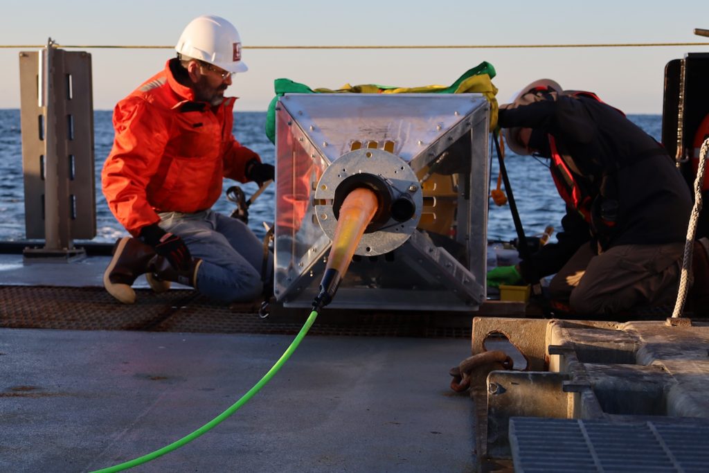 Two engineers wearing hard hats and orange life vests prepare a scientific instrument for deployment. The engineers are crouched down, kneeling on the wet, silver metal deck of a research ship. The instrument in between them has a square, silver metal frame with a green cable extending towards the camera, laying flat on the ship deck. In the foreground are assorted maritime equipment. In the background is blue ocean with hazy, orangish-blue sky on the horizon.