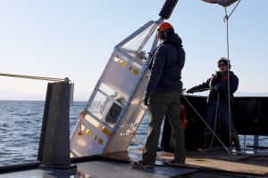 Two members of MBARI’s marine operations crew deploy a scientific instrument from the back deck of a research ship on a bright sunny day. The crew are wearing hard hats and orange life vests. The instrument has a silver metal frame with white plastic shrouds, with two yellow plastic spheres and one silver metal sphere inside. The instrument is suspended from a metal crane outside the frame. In the foreground is the silver metal deck of a research ship. In the background are blue ocean, coastal mountains, and hazy blue sky.