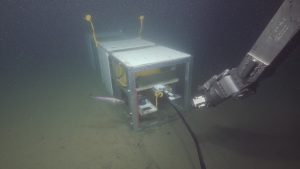 A scientific instrument with a rectangular silver metal frame, white plastic panels, and a yellow plastic sphere rests on the muddy seafloor. A black cable connected to the instrument extends off-screen toward the camera. In the foreground are the silver metal robotic arm of a deep-sea submersible and a silver fish. The background is greenish-brown mud.