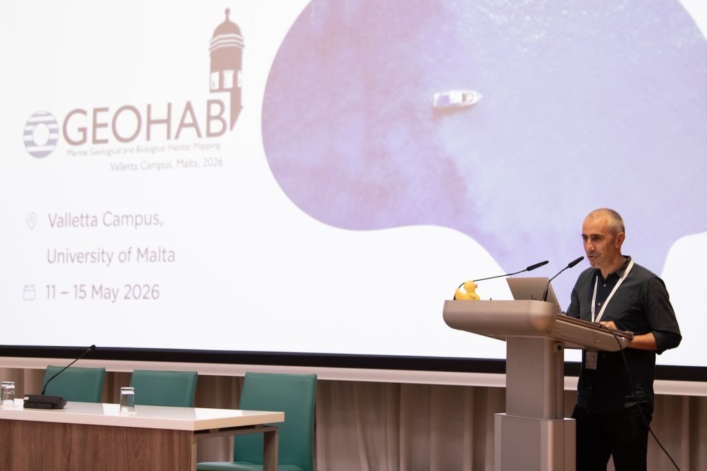 A scientist standing behind a silver podium presents at a scientific conference. In the background is a presentation slide projected on a large screen. The slide has the conference logo artwork and reads, GEOHAB, Valletta Campus, University of Malta, 11 – 15 May 2026.