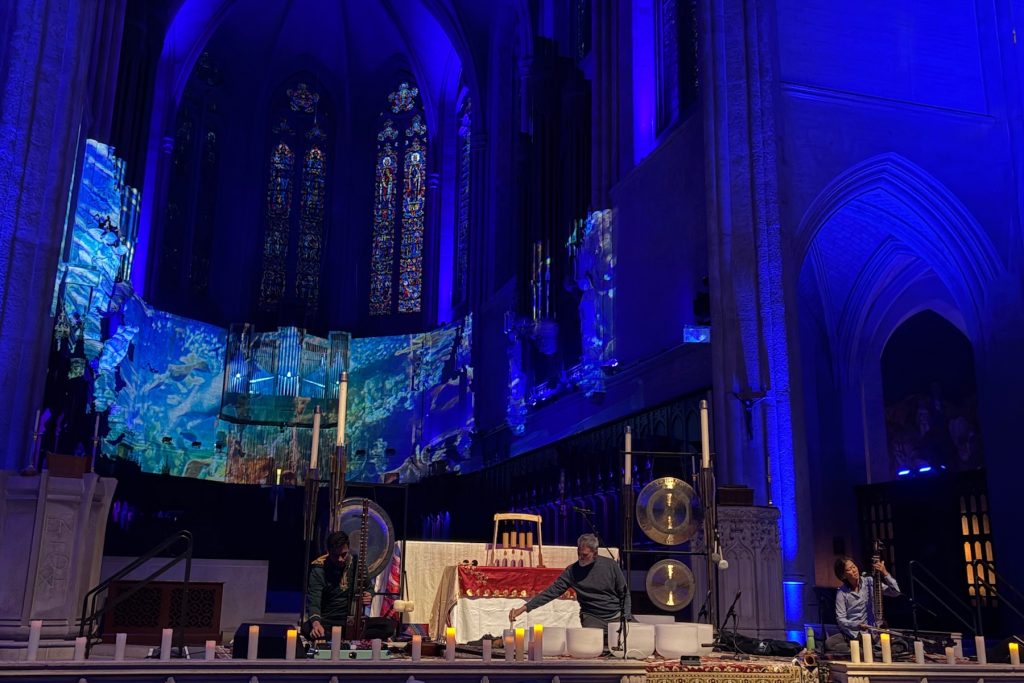 Three musicians perform on the stone altar of a historic gothic cathedral. The musicians are seated and playing musical instruments under a warm spotlight. In the foreground are white candles. In the background is the stone nave of the cathedral, with stained-glass windows and projected underwater video.