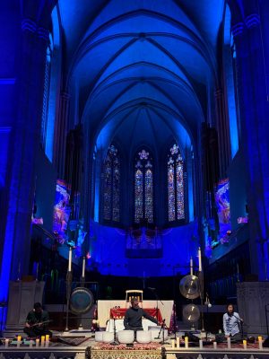 Three musicians perform on the stone altar of a historic gothic cathedral. The musicians are seated and playing musical instruments under a warm spotlight. In the foreground are white candles. In the background is the soaring stone nave of the cathedral, with stained-glass windows and projected underwater video.