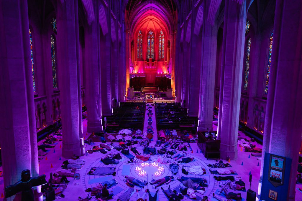 Sound bath participants lying on yoga mats and sitting in wooden pews of Grace Cathedral. The cathedral’s stone pillars are illuminated in purple light and the cathedral’s altar is illuminated in yellow and red lights.