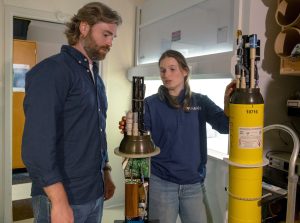 Two researchers examine chemical sensors on robotic floats. The researcher on the left has brown hair and a brown beard, and is wearing a blue button-down shirt and blue jeans. The researcher on the right has shoulder-length brown hair and is wearing a navy blue pullover with the MBARI logo and blue jeans. In between the two researchers is a sensor instrument with a black plastic housing and a green computer motherboard. On the right is a robotic float with a yellow plastic housing. The background is a science lab with white walls, wooden shelves, and wooden cabinets.