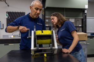 Two scientists inspect a roll of thin yellow fiber-optic cable on a large black plastic spool on the counter inside a brightly lit scientific lab.