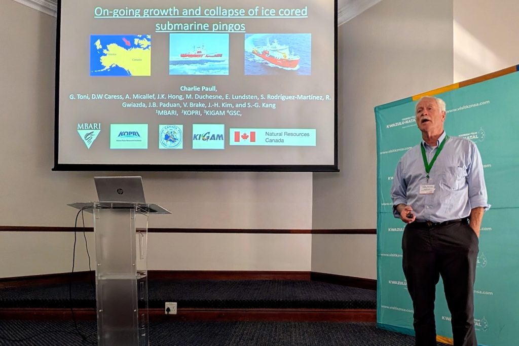 A scientist presents a slide at a scientific conference. He is standing on a raised stage to the right of a screen displaying a black slide with multi-colored photos and institutional logos titled, On-going growth and collapse of ice cored submarine pingos. In the background are a teal banner, a clear plastic podium with a silver laptop computer, and the white walls of a hotel ballroom.