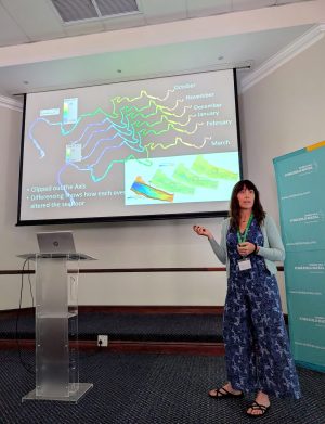 A scientist presents a slide at a scientific conference. She is standing on a raised stage to the right of a screen displaying multi-colored plats of scientific data. In the background are a teal banner, a clear plastic podium with a silver laptop computer, and the white walls of a hotel ballroom.