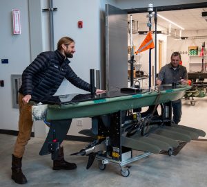 Two engineers maneuver a cart transporting a dark-green ocean robot. The engineer in the foreground with a beard and long brown hair is wearing a puffy black jacket, khaki pants, and brown boots. The engineer in the background with short gray hair is wearing a gray short-sleeved shirt and blue jeans. The robot has a canoe-shaped top with two tall poles and an orange flag, with a black propeller and six gray-green fin-shaped slats underneath. The robot is on a cart with a silver base and four small black wheels. The engineers are moving the robot from a high bay with white walls and a gray concrete floor through a set of wide silver metal doors into a lab with white walls, a white laminate floor, and oceanographic research equipment.
