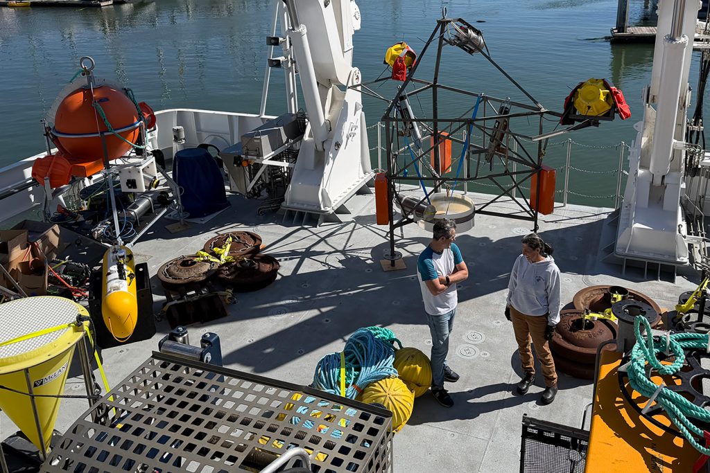 Two researchers stand on the back deck of a ship that is loaded with large oceanic instruments, including yellow sediment traps, an instrument with a large rectangular orange float, spherical orange and yellow devices, and the ship’s A-frame.