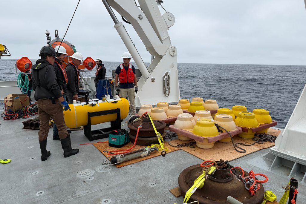 A group of five crew members in hard hats and life vests stands on a ship's deck near a yellow torpedo-shaped instrument and pallets of flotation spheres, preparing for a deployment beside a large white A-frame crane.