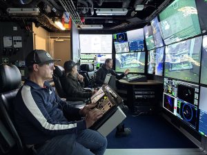 Three people sit at control consoles in a dark submersible control room, monitoring a wall of screens showing underwater video, navigation data, and vehicle telemetry.