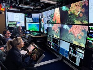 Three researchers control a robotic submersible from a dimly lit control room aboard a research ship, viewing live video feeds of a deep-sea coral community displayed on large screens alongside navigation and sonar data.