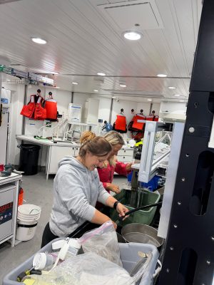 Two smiling researchers work together in a shipboard lab, sieving a sample in a sink. A tub of sampling supplies is in the foreground, and orange life vests hang on white walls in the background.