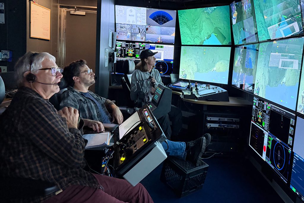 Three people sit in a dim submersible control room and monitor a bank of screens showing live video feed of the seafloor, maps, and data streams.