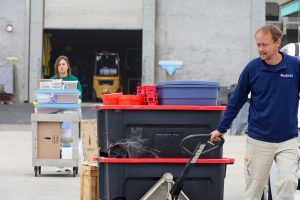 A man in an MBARI pullover pulls a hand truck loaded with storage bins labeled Wood Collection Rings across a loading area, while a woman pushes a rolling cart of supply boxes in the background near an open warehouse door.