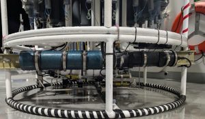A close-up photo shows instruments on the round white metal frame of a scientific instrument. On the left is a blue metal cylinder housing an oceanographic sensor, and on the right is a black metal cylinder housing a smaller oceanographic sensor. Above is a carousel of gray plastic sampling bottles. In the background is the wet gray metal deck of a research ship.