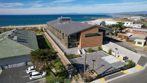 A photo from an aerial drone shows the southeast corner of MBARI’s new robotic technology lab. The lab has wood panel siding on the east wall and gray cement siding on the south wall. To the left, is the green roof of an adjacent MBARI building. To the right, are other buildings in Moss Landing, California. An empty gray parking lot is in the foreground, and the blue ocean and hazy blue sky with gray clouds are in the background.