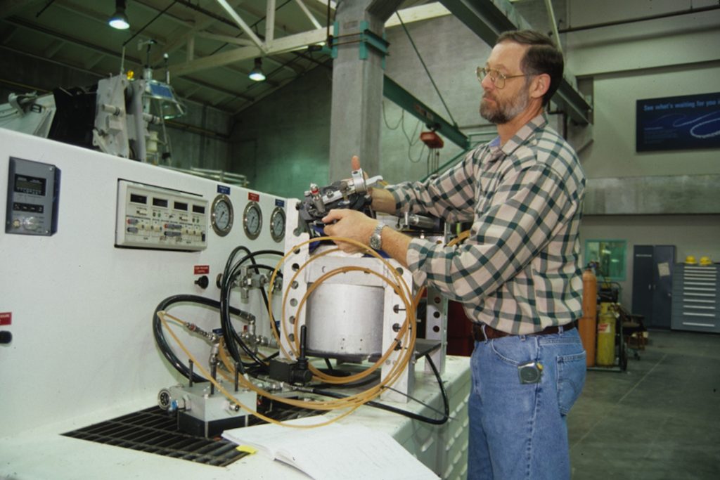 An engineer prepares scientific equipment on a counter in a machine shop. In the background are gray concrete walls and assorted engineering equipment.