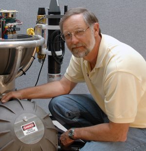 An engineer crouches down for a photo with a disassembled scientific instrument inside a silver metal sphere. In the background is a textured gray wall.