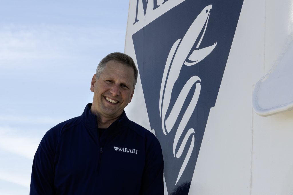 MBARI’s Director of Marine Operations with short gray hair wearing a blue pullover with the MBARI logo leans against the exterior of an MBARI research vessel. The ship’s metal exterior is white metal, with a dark-blue triangle and the white MBARI gulper eel logo. The background is blue sky with wispy white clouds.