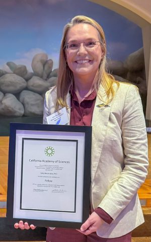A scientist holding a framed award certificate poses for a photo inside a science museum.