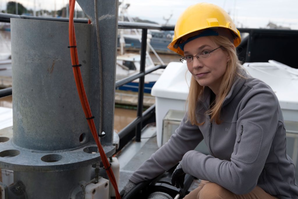 A scientist wearing a yellow hard hat poses for a photo while crouched down next to a scientific instrument on the outdoor deck of a research ship on an overcast day.