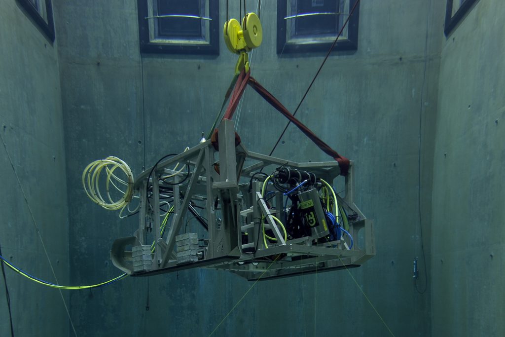 The seafloor mapping toolsled with a weathered metal frame, silver and black metal canisters, and a tangle of green, blue, and yellow cables. The toolsled was photographed while submerged in MBARI’s test tank and suspended by a yellow crane hook and red ropes as the lidar canister projects three green lasers toward the bottom of the tank. Three gray, concrete walls are visible in the background.