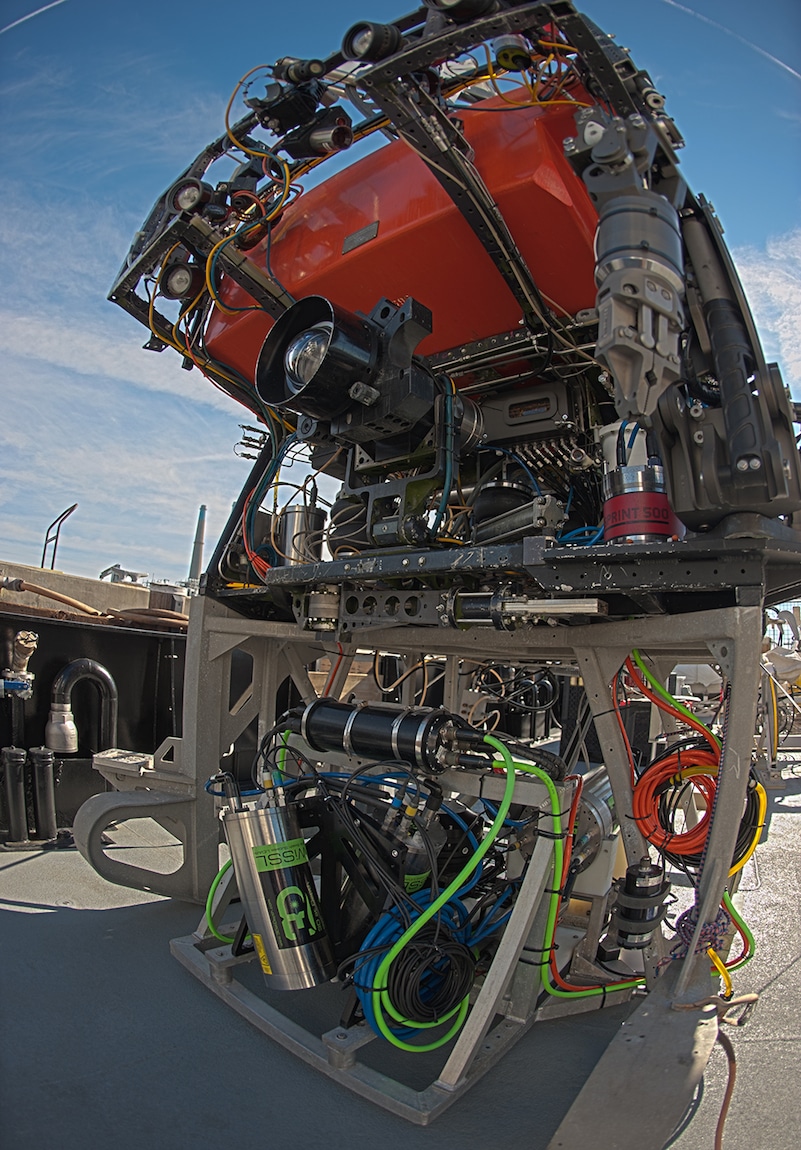 Metal frame with seafloor mapping toolsled with silver and black canisters and bright green cables installed below a robotic submersible with a black camera, metal manipulator arm, and bright orange float. Photographed with a fisheye lens on the metal deck of a research vessel with blue sky and clouds in the background.