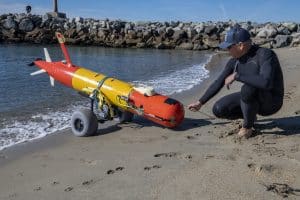 An MBARI engineer wearing a black wetsuit inspects an orange-and-yellow-colored, torpedo-shaped, autonomous robot on the sandy beach with a rocky jetty in the background.