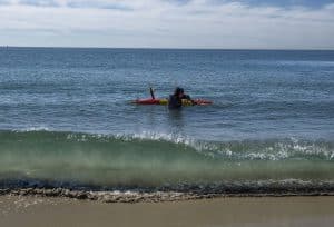 Two MBARI engineers wearing black wetsuits secure an orange-and-yellow-colored autonomous robot in the surf as waves crash on a sandy beach.