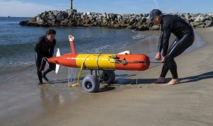 Two MBARI engineers in black wetsuits recover an orange-and-yellow-colored, torpedo-shaped, autonomous robot using a black cart with large, gray wheels on the wet, sandy beach with a rocky jetty in the background.