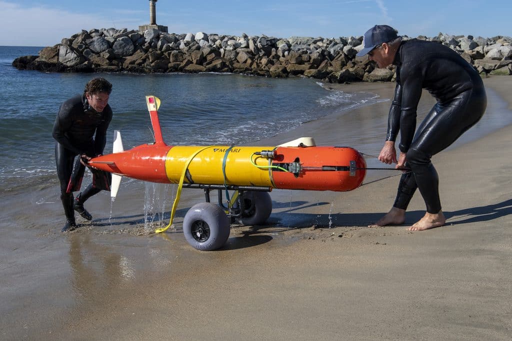 Two MBARI engineers in black wetsuits recover an orange-and-yellow-colored, torpedo-shaped, autonomous robot using a black cart with large, gray wheels on the wet, sandy beach with a rocky jetty in the background.