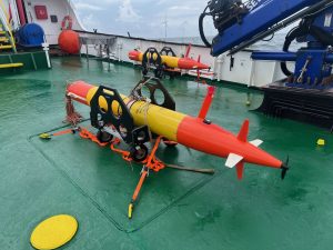 Two tubular underwater robots sit secured in black carts in preparation for deployment. The robots have a yellow body with an orange nose cone and orange tail. They are strapped into black carts on the green metal deck of the research ship. Various maritime equipment is in the background. In the distance is overcast gray sky, grayish-blue ocean, and white metal wind turbines.