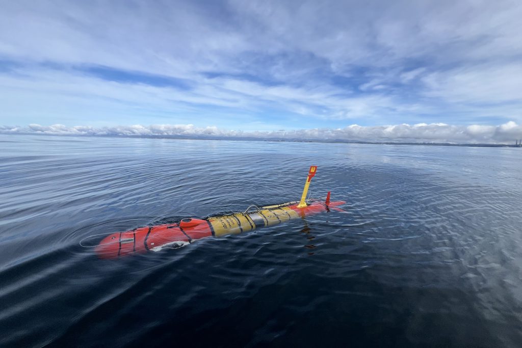 A torpedo-shaped, orange-and-yellow underwater robot glides at the ocean’s surface. The robot’s tail “fin” extends above the water’s surface. This robot was photographed during a deployment on a cloudy day, with dark blue, rippled ocean in the foreground, billowy gray clouds on the horizon, and blue sky with hazy gray clouds above.