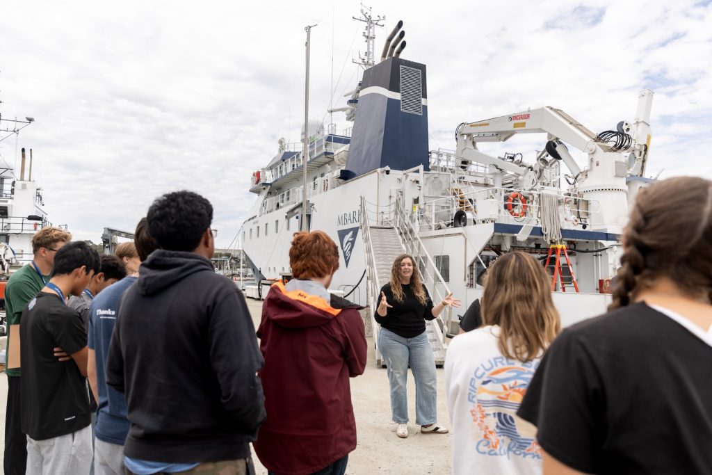An MBARI researcher speaks with a large group of high school students outdoors on MBARI’s dock. She is standing in front of two scientific research ships. In the background is cloudy blue sky.